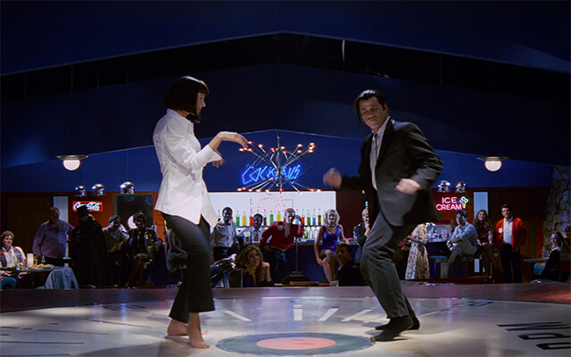 Vincent Vega e Mia Wallace dançando na clássica cena de Pulp Fiction em um palco com temática retrô.
