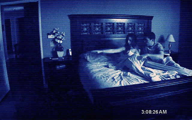 Casal sentado na cama em imagem escura de câmera de segurança, do filme Atividade Paranormal.